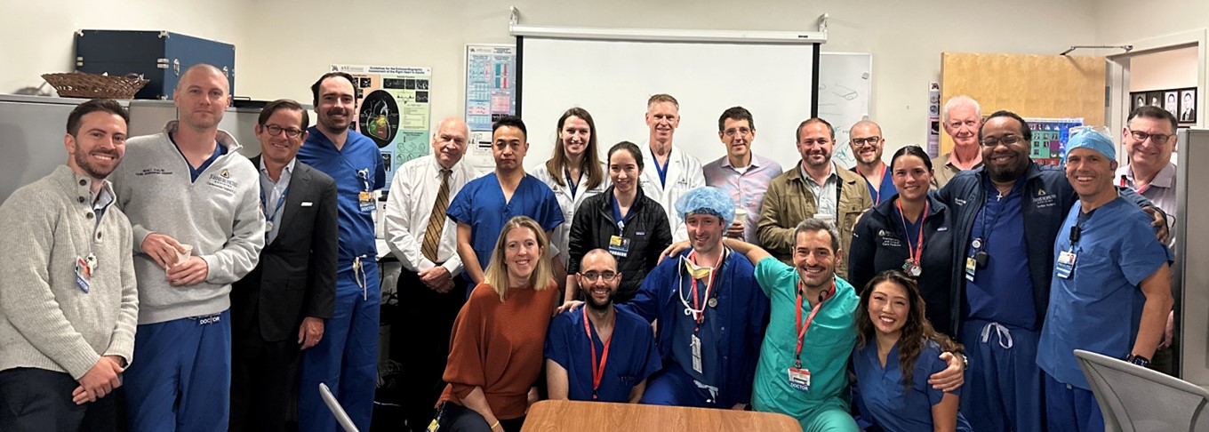 A large group of approximately 20 medical professionals, including the six new cardiothoracic anesthesia fellows and two cardiac surgery residents, posing for a group photo in a conference room. The team is dressed in a mix of blue surgical scrubs, white medical lab coats, and professional business attire. They are gathered around a wooden conference table, with some individuals holding coffee cups. The background includes a white projector screen and anatomical heart posters on the wall.