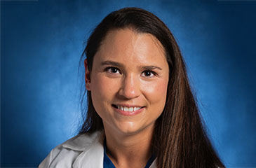 A professional headshot of Dr. Chelsey Santino, MD, an Assistant Professor of Anesthesiology and Critical Care Medicine. She has long brown hair and is smiling warmly, wearing a white medical lab coat over a blue surgical scrub top. The portrait is set against a textured blue studio background.