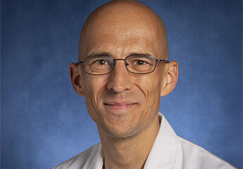 A professional headshot of Dr. Tymoteusz (Tym) Kajstura, MD, an Assistant Professor of Anesthesiology and Critical Care Medicine. He is a man with glasses and a friendly smile, wearing a white medical lab coat over a blue surgical scrub top. The portrait is set against a solid blue studio background.