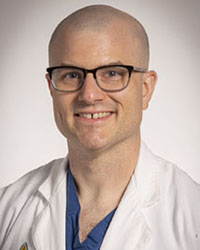 Dr. Joseph Walpole, recipient of the 2025 Outstanding Resident Research Award, smiling while holding his award certificate in a modern hospital atrium. He is wearing blue surgical scrubs and a white medical lab coat.