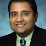 A professional headshot of Dr. Shreosi (Sam) Das, PhD, Assistant Professor of Anesthesiology and Critical Care Medicine. He is smiling warmly and wearing a black blazer over a pale green button-down shirt with a blue patterned tie against a textured blue-green studio background.