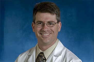 A professional headshot of Dr. Adam Schiavi, MD, PhD, an Associate Professor in Anesthesiology and Critical Care Medicine. He is smiling and wearing glasses, a tan dress shirt, a patterned tie, and a white Johns Hopkins medical lab coat with his name and departments embroidered in blue on the chest. The portrait is set against a textured blue studio background.