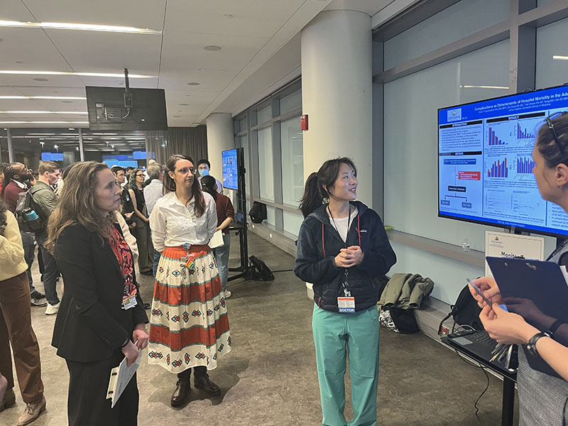A group of researchers and faculty members engaged in a discussion in front of a digital poster titled "Complications as Determinants of Hospital Mortality in the Adult ECMO Population" during the Quality Improvement session at ACCM Research Day.