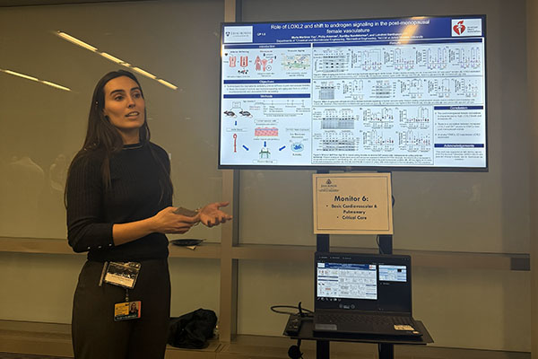 A researcher standing by a digital monitor and presenting her scientific poster titled "Role of LOXL2 and shift to androgen signaling in the post-menopausal female vasculature" at the Basic Cardiovascular & Pulmonary poster session.