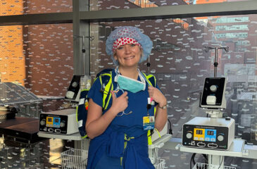 A portrait of female CRNA Paige McMurtrie smiling in a clinical workspace. She is wearing blue surgical scrubs, a stethoscope, and a blue surgical cap with a heart-patterned headband. She stands in front of a large window overlooking brick hospital buildings, flanked by medical monitoring equipment and electrosurgical generators.