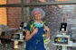A portrait of female CRNA Paige McMurtrie smiling in a clinical workspace. She is wearing blue surgical scrubs, a stethoscope, and a blue surgical cap with a heart-patterned headband. She stands in front of a large window overlooking brick hospital buildings, flanked by medical monitoring equipment and electrosurgical generators.