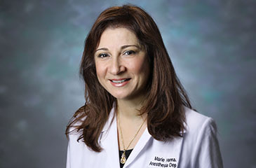A professional headshot of Dr. Marie Hanna, MD, MEHP, Professor of Anesthesiology and Critical Care Medicine. She is smiling and wearing a white medical lab coat with "Marie Hanna, Anesthesia Dep" embroidered on the left side, over a black top and a gold pendant necklace. The portrait is set against a textured teal and blue studio background, typical of Johns Hopkins Medicine faculty photography.