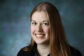 A professional headshot of Dr. Christina Miller, MD, an Associate Professor of Anesthesiology and Critical Care Medicine and Otolaryngology-Head and Neck Surgery. She is smiling warmly with a bright, engaged expression against a textured, professional blue studio background. She is wearing a black collared button-down shirt. The photograph has a high-fidelity, polished finish.
