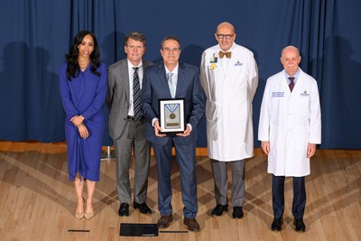 Group portrait taken on a stage with a blue curtain backdrop, showing a Johns Hopkins Medicine clinical leader in a white lab coat smiling while holding a "Clinical Excellence" medal award plaque. They are standing center-stage flanked by four other Johns Hopkins institutional and clinical leaders, including Dean/CEO Theodore DeWeese and President Kevin Sowers, who are present to recognize the achievement.