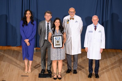 Group portrait taken on a stage with a blue curtain backdrop, showing a Johns Hopkins Medicine clinical leader in a white lab coat smiling while holding a "Clinical Excellence" medal award plaque. They are standing center-stage flanked by four other Johns Hopkins institutional and clinical leaders, including Dean/CEO Theodore DeWeese and President Kevin Sowers, who are present to recognize the achievement.
