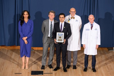 Group portrait taken on a stage with a blue curtain backdrop, showing a Johns Hopkins Medicine clinical leader in a white lab coat smiling while holding a "Clinical Excellence" medal award plaque. They are standing center-stage flanked by four other Johns Hopkins institutional and clinical leaders, including Dean/CEO Theodore DeWeese and President Kevin Sowers, who are present to recognize the achievement.
