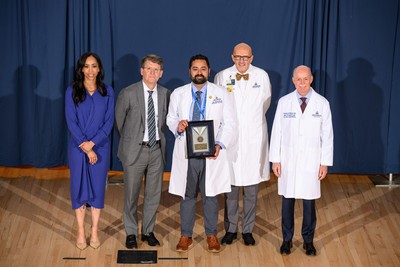 Group portrait taken on a stage with a blue curtain backdrop, showing a Johns Hopkins Medicine clinical leader in a white lab coat smiling while holding a "Clinical Excellence" medal award plaque. They are standing center-stage flanked by four other Johns Hopkins institutional and clinical leaders, including Dean/CEO Theodore DeWeese and President Kevin Sowers, who are present to recognize the achievement.