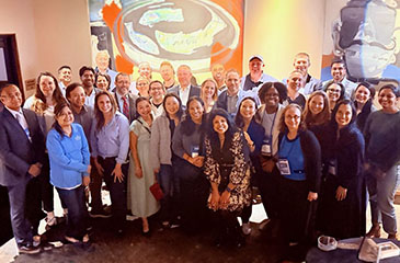 A large group photo of approximately 30 members of the Johns Hopkins Pediatric Anesthesia and Pain Medicine teams gathered in an event space. The team, including faculty and fellows, is dressed in professional and business-casual attire. Several individuals are wearing blue conference lanyards. They are posing in front of large, colorful contemporary paintings, one featuring fish and another a stylized face.