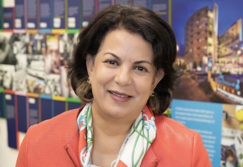 Professional portrait of Dr. Anna Varughese, wearing an orange blazer and a patterned scarf, smiling in front of a colorful wall display of medical milestones.