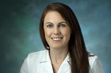 A professional headshot of Dr. Caitlin O'Brien, MD, an Assistant Professor of Anesthesiology and Critical Care Medicine. She is smiling warmly and wearing a clean white medical lab coat against a textured teal-blue studio background.