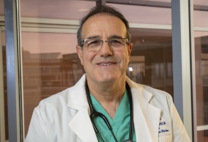 Professional headshot of Dr. Ozan Akca, wearing a white lab coat over green scrubs with a stethoscope around his neck, smiling against a windowed background.