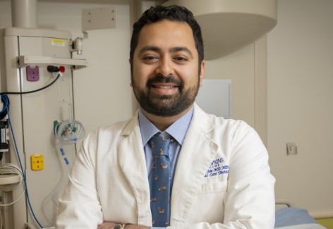 Professional headshot of Dr. Rohan Mathur, wearing a white Johns Hopkins lab coat and a blue patterned tie, smiling in front of clinical equipment.