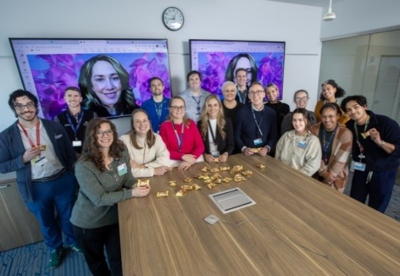 A large group of diverse clinical and research staff, known as the S’Mores team, gather around a wooden conference table covered in snacks. Two large monitors in the background show a remote colleague joining via video call.
